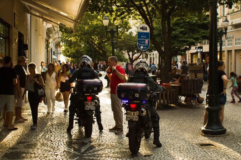 Guardas municipais em motocicletas patrulham uma rua movimentada, com pedestres ao redor.