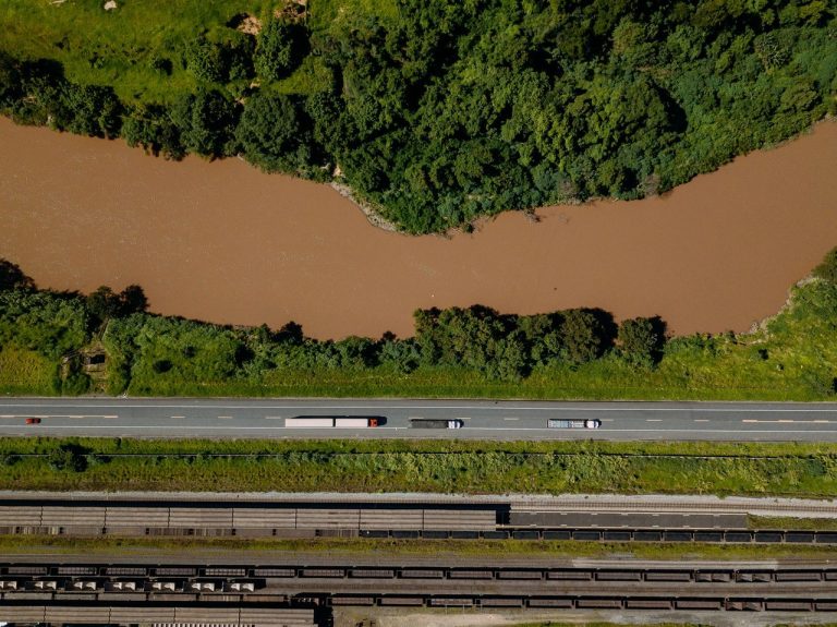 Vista aérea de caminhões circulando por uma rodovia brasileira às margens de um rio.
