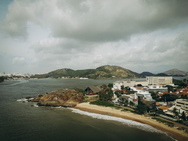 Serra lidera turismo capixaba com 88,1% de visitantes de outros estados Vista aérea de uma praia junto a uma área urbana no Espírito Santo, cercada por morros e sob céu nublado.