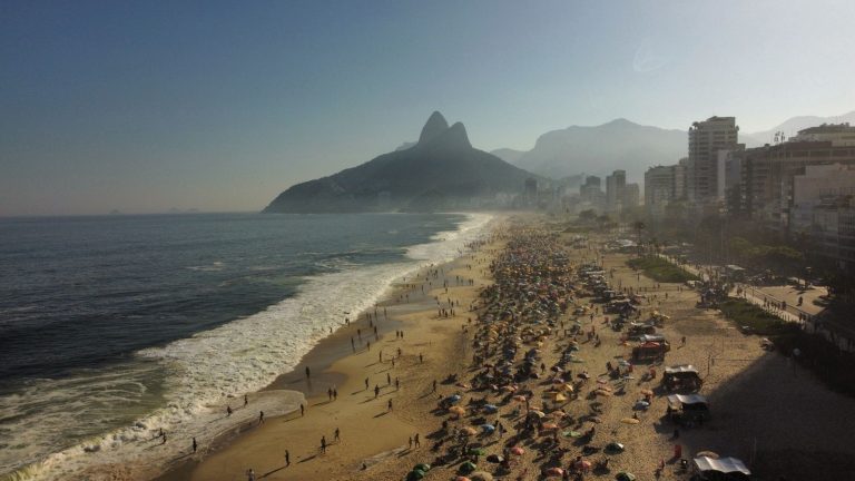 Turismo injeta R$ 22,5 bilhões na economia do Rio até agosto de 2026 Praia de Ipanema movimentada, com montanhas e a paisagem urbana do Rio de Janeiro ao fundo.